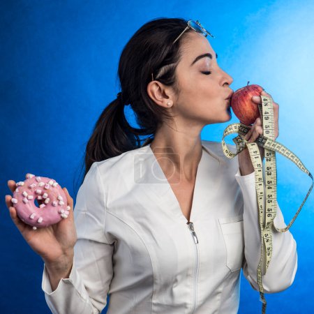 Foto de Dietista en bata blanca con el pelo recogido sostiene en una mano un dulce mientras besa una manzana que sostiene en la otra mano, aislada sobre fondo azul - Imagen libre de derechos