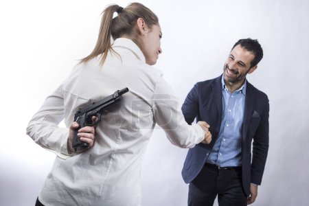 Foto de Chica rubia con camisa blanca sostiene un arma en su mano mientras se da la mano con un hombre de pelo oscuro vestido con chaqueta y camisa, aislado sobre fondo blanco - Imagen libre de derechos