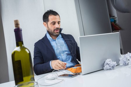 Foto de Gerente de cabello oscuro vestido con un traje y molesto y sentado en su oficina y sosteniendo una botella de vino cerca de su computadora - Imagen libre de derechos