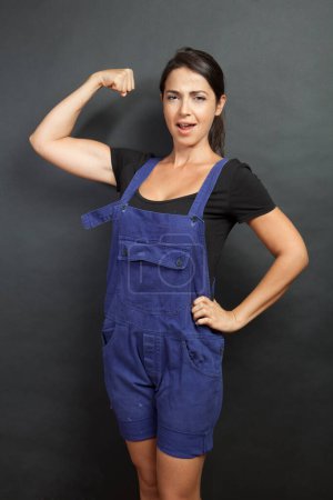 Photo for Forced white girl with black hair and work overalls shows the arm muscles, isolated on black background - Royalty Free Image