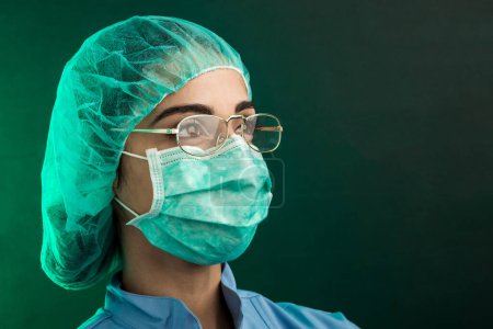 Téléchargez les photos : Médecin en blouse bleue, bonnet opératoire vert et masque chirurgical facial semble grave à travers ses lunettes, isolé sur fond vert - en image libre de droit