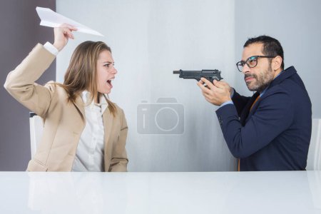 Photo for Two colleagues sitting in their workplace, the manager points the gun at the surprised secretary who launches a paper plane - Royalty Free Image