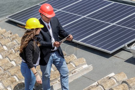 Foto de Pareja de ingenieros con casco protector comprueban el estado de un sistema fotovoltaico en el techo de una casa utilizando una tableta de ingenieros sistema fotovoltaico - Imagen libre de derechos