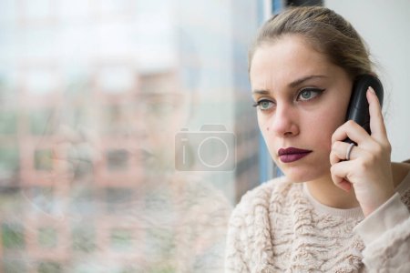 Foto de Hermoso retrato de chica rubia con ojos verdes de pie cerca de una ventana de vidrio dentro de un edificio mientras habla por teléfono - Imagen libre de derechos