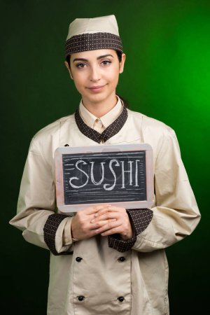 Photo for European cook in uniform holds a blackboard with Sushi written on it, isolated on a green background. - Royalty Free Image