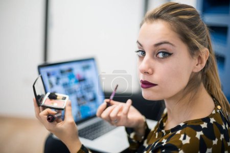 Photo for Young woman dressed casual finishes makeup in her office while putting on lipstick while standing - Royalty Free Image