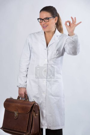 Téléchargez les photos : Blonde femme médecin avec des lunettes et les cheveux ramassés, tient un sac de médecin et fait le signe ok, isolé sur fond blanc - en image libre de droit