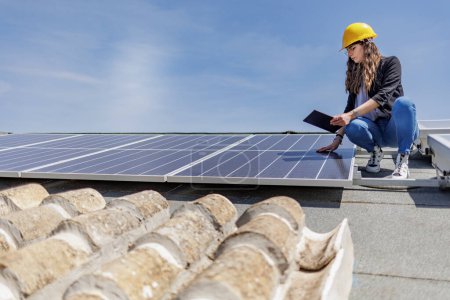 Téléchargez les photos : Une ingénieure portant un casque jaune vérifie l'état d'un système photovoltaïque sur le toit d'une maison avec une tablette à la main - en image libre de droit