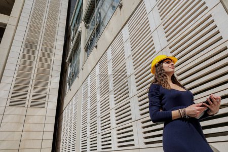 Téléchargez les photos : Femme élégamment habillée avec un casque de protection jaune, tenant sa tablette à la main dans un contexte urbain moderne - en image libre de droit