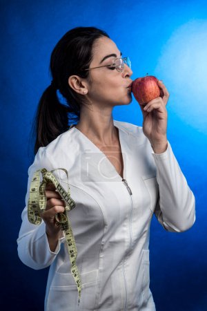 Foto de Dietista en bata blanca con el pelo recogido besa una manzana que sostiene en la otra mano, aislada sobre fondo azul - Imagen libre de derechos