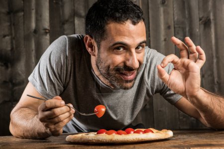 Foto de Hombre de pelo oscuro con barba está a punto de comer una pizza con tomates cherry, aislado sobre fondo de madera - Imagen libre de derechos