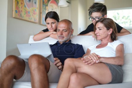 Photo for Family made up of 4 people is concentrated looking at the tablet in a moment of relaxation in the living room at home - Royalty Free Image