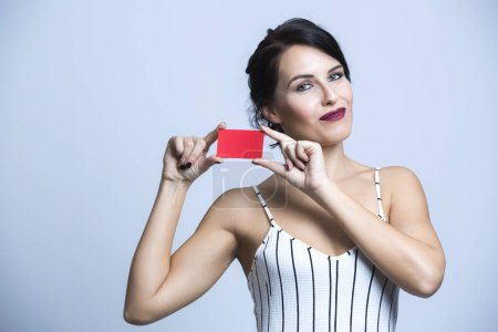 Photo for White woman with black hair dressed in a white striped jumpsuit points to a red card, isolated on white background - Royalty Free Image