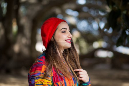 Foto de Retrato de hermosa chica blanca en suéter colorido y gorra roja, aislado sobre el fondo de la naturaleza mirando a la distancia - Imagen libre de derechos
