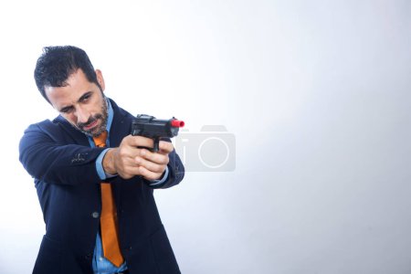 Foto de Hombre de pelo oscuro con barba vestido de traje con chaqueta naranja y corbata apunta un arma en blanco - Imagen libre de derechos