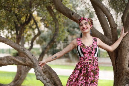 Photo for Beautiful smiling girl with red lipstick dressed in a colorful dress and a headband of the same material, stands in the middle of a natural park - Royalty Free Image
