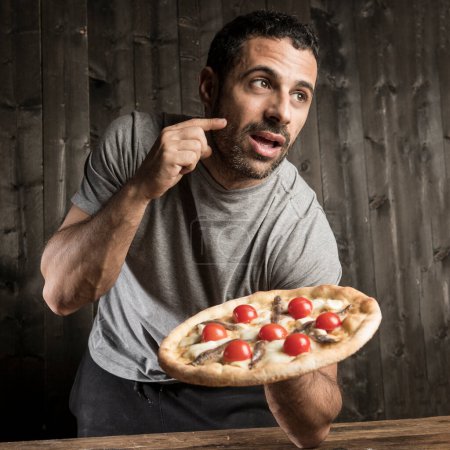 Téléchargez les photos : Un homme aux cheveux bruns barbu est sur le point de manger une pizza aux tomates cerises, isolé sur fond de bois - en image libre de droit