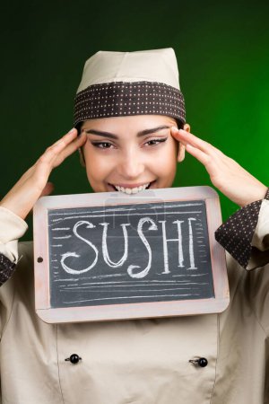 Téléchargez les photos : Cuisinier européen en uniforme tient un tableau noir avec Sushi écrit dessus, isolé sur un fond vert. - en image libre de droit