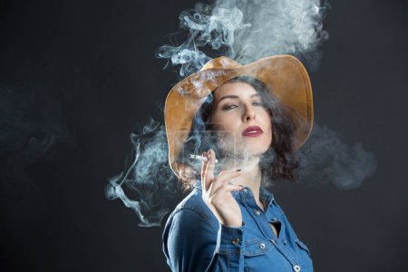 Foto de Chica morena con camisa vaquera y sombrero de vaquero, fuma un cigarrillo produciendo mucho humo, aislado sobre fondo gris - Imagen libre de derechos