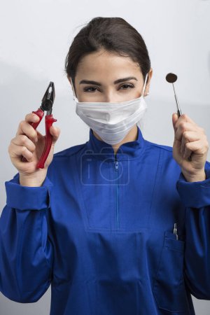 Foto de Dentista femenina con máscara quirúrgica y capa azul muestra herramientas de trabajo en una mano y alicates de bricolaje en la otra, aislados sobre fondo azul claro - Imagen libre de derechos