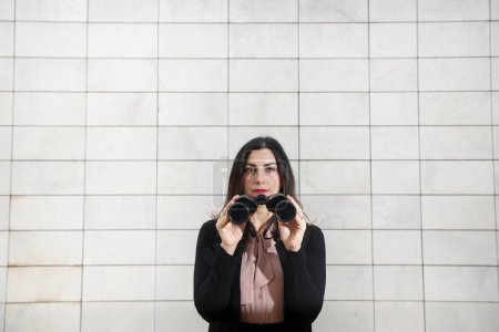 Photo for Brunette female manager, dressed in black business suit, holds binoculars in hand to look away, isolated on white rectangular tile wall background - Royalty Free Image
