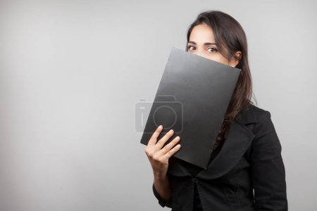 Photo for White girl with dark hair dressed in black jacket holds a black folder in hand and looks puzzled isolated on white background - Royalty Free Image