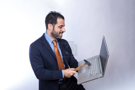 Téléchargez les photos : Homme aux cheveux bruns avec barbe, vêtu d'un costume chemise, veste et cravate orange, prend un ordinateur portable dans sa main avec un marteau, isolé sur fond blanc - en image libre de droit