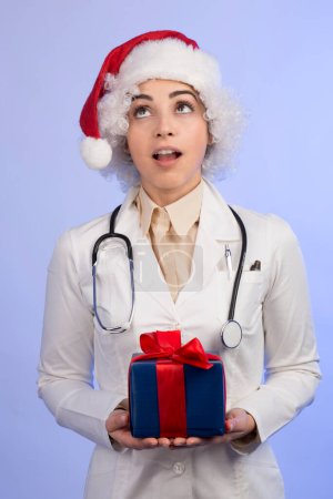 Téléchargez les photos : Femme médecin en manteau blanc et chapeau santa claus détient un cadeau en cadeau, isolé sur fond clair - en image libre de droit