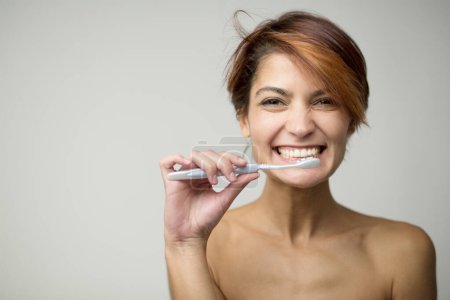 Téléchargez les photos : Belle fille aux cheveux courts se brosse les dents avec une brosse à dents, isolée sur fond clair - en image libre de droit