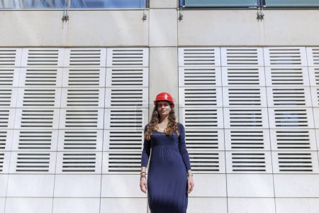 Foto de Mujer elegantemente vestida con casco protector rojo en una posición estática se ve serio en la distancia en un entorno urbano moderno - Imagen libre de derechos