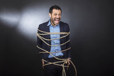 Foto de Hombre en traje desesperado grita atado con una cuerda sobre una silla aislada sobre un fondo negro - Imagen libre de derechos