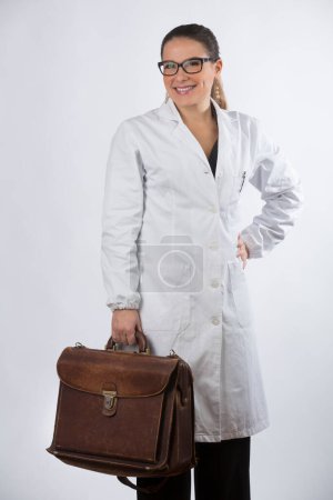 Photo for Blonde female doctor with eyeglasses and gathered hair, holds a doctor's bag and makes the ok sign, isolated on white background - Royalty Free Image