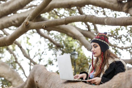 Téléchargez les photos : Brunette fille habillée en noir avec un casque sur la tête utilise son ordinateur portable appuyé sur un arbre dans un parc - en image libre de droit