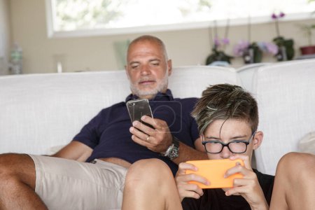 Téléchargez les photos : Père et fils assis dans le salon à la maison s'isolent avec des jeux électroniques et des téléphones mobiles - en image libre de droit