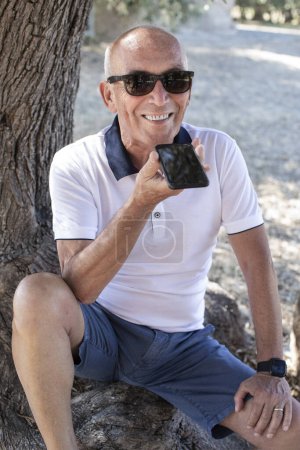 Téléchargez les photos : Portrait d'un bel homme mature dans le parc avec smartphone en plein air - en image libre de droit