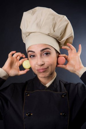 Photo for Chef woman in uniform with chef hat jokes with a pair of macarons isolated on black background - Royalty Free Image