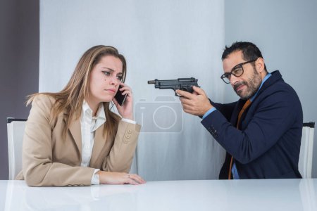Foto de Dos colegas sentados en su lugar de trabajo, el gerente apunta el arma a la secretaria aburrida que habla por teléfono - Imagen libre de derechos
