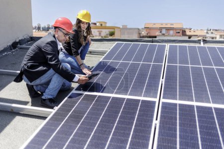 Foto de Un par de ingenieros con cascos protectores comprueban el estado de un sistema fotovoltaico en el techo de una casa usando una tableta - Imagen libre de derechos