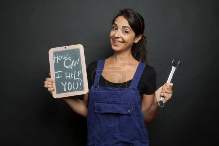 Photo for Mechanic woman in blue overalls holding a mechanic's tool and a blackboard with "how can I help you?", isolated on black background - Royalty Free Image