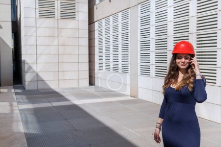Foto de Mujer elegantemente vestida de rojo sombrero duro habla con expresión seria en el teléfono en el entorno urbano moderno - Imagen libre de derechos