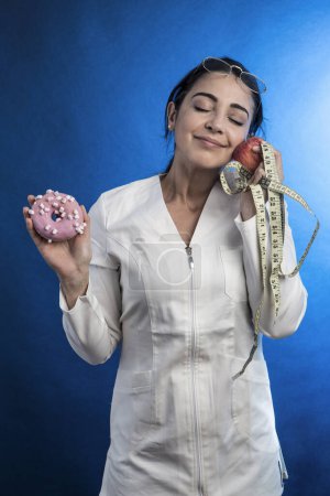 Foto de Médico con una bata blanca y sostiene un pastel en una mano y una manzana en la otra a la que muestra mucho afecto, aislado sobre fondo azul - Imagen libre de derechos