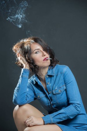 Foto de Chica morena con camisa vaquera y sombrero de vaquero, fuma un cigarrillo produciendo mucho humo, aislado sobre fondo gris - Imagen libre de derechos