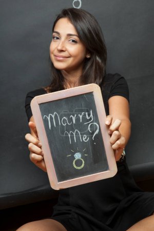 Téléchargez les photos : Une femme aux cheveux bruns vêtue de noir tient un tableau noir avec "Épouse-moi ?" écrit dessus isolé sur fond noir avec le dessin d'un roman de dessin animé - en image libre de droit