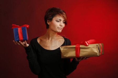 Téléchargez les photos : Belle fille avec chemise noire et heureuse d'avoir deux cadeaux à déballer dans ses mains, isolée sur fond rouge - en image libre de droit