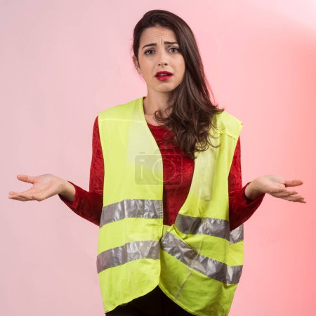 Photo for White girl with dark hair and a yellow emergency vest on her is desperate after committing an accident, isolated on pink background - Royalty Free Image