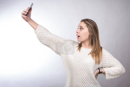 Téléchargez les photos : Blonde fille avec chemise blanche prend un selfie avec téléphone mobile, isolé sur fond blanc - en image libre de droit