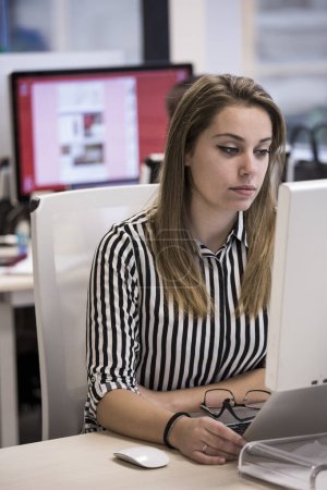 Téléchargez les photos : Gérant blonde avec des lunettes et chemise rayée travaille dans le bureau assis devant son lieu de travail - en image libre de droit