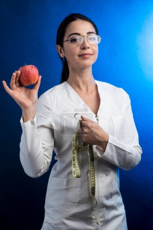 Foto de Dietista en bata de laboratorio con el pelo recogido sostiene en una mano una manzana y una cinta métrica, aisladas sobre un fondo azul - Imagen libre de derechos