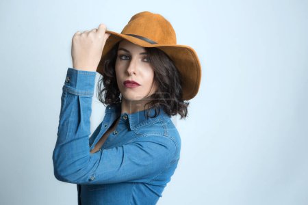 Photo for White model with black hair dressed in a denim shirt and a brown hat, isolated on dark background - Royalty Free Image