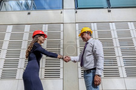 Foto de Par de ingenieros con cascos de protección estrechando la mano de acuerdo en un contexto de arquitectura moderna - Imagen libre de derechos